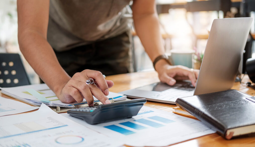 Close up of businessman or accountant hand holding pen working on calculator to calculate business data, accountancy document and laptop computer at office, business concept