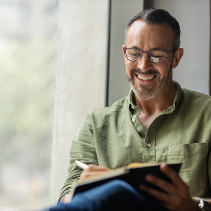 Mature man with grey and black beard and glasses smiling, sitting near a bright sunny window writing in notebook with copy space