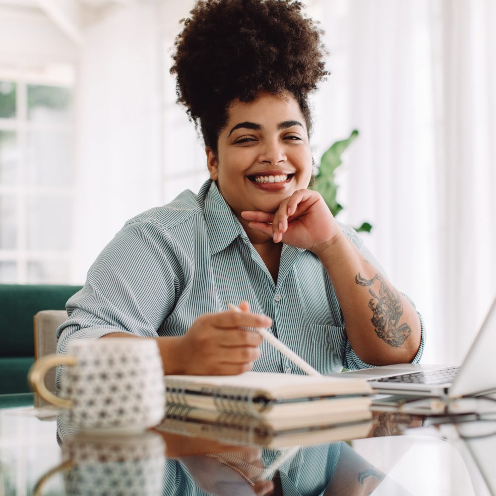 Female sitting at desk with laptop and diary. Woman working from home office looking at camera and smiling.