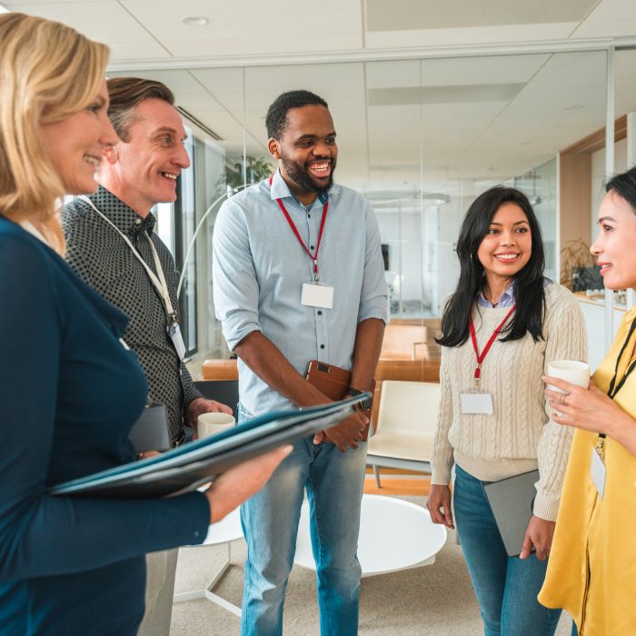 3/4 length image of a multiracial business team of mid adult people in a conference room, all wearing casual clothes and name tags while chatting and smiling to each other, standing in a meeting room.
