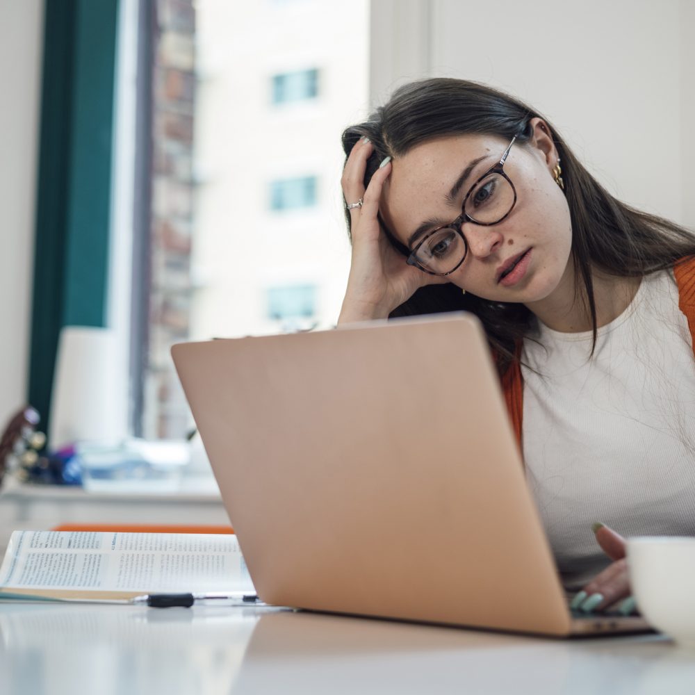 Front view of a young woman wearing casual clothing sitting at a desk using a laptop to study, she is a university student. She is feeling stressed and she has her head in her hand.