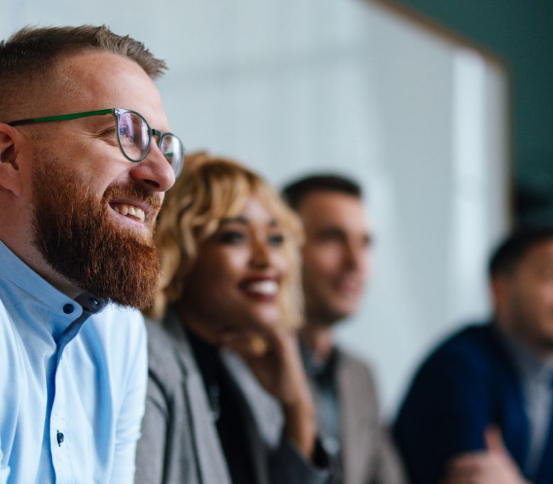 Smiling businessman and unrecognizable multi-ethnic employees sitting in a row at their company desk and listening to their boss.