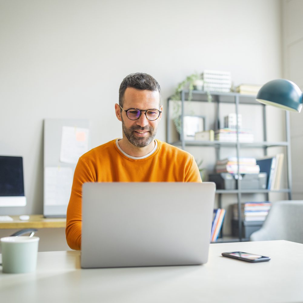 Businessman wearing casual clothing and eyeglasses sitting at his desk and using laptop in the office.
