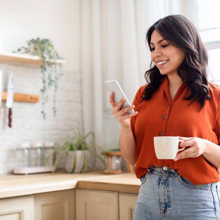 Smiling young woman enjoying her coffee while using her smartphone in a homey kitchen setting, copy space