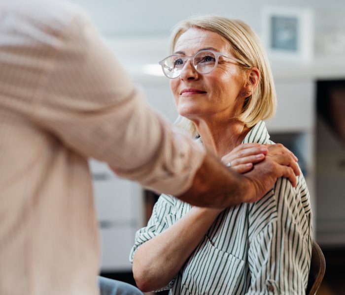 Senior woman being emotionally supported by a male friend indoors. The photo showcases warmth, care, and friendship in a modern home environment.