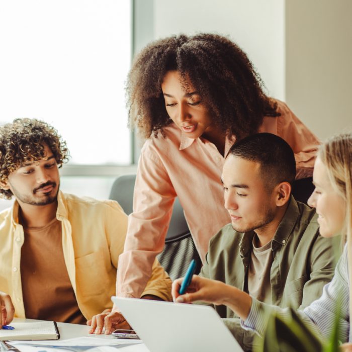 Diverse team of young professionals are collaborating on a project, using a laptop and going over paperwork in a bright office