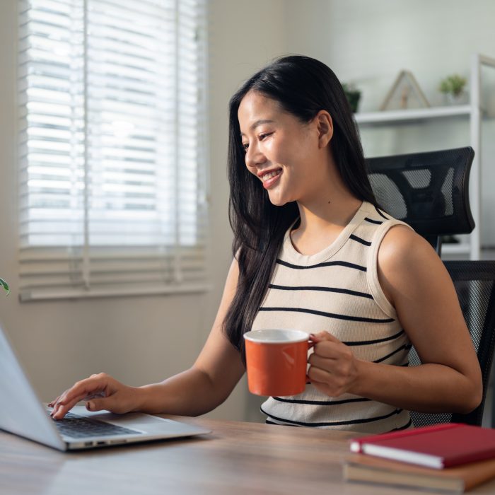A confident, satisfied Asian businesswoman is enjoying her morning coffee while working on her laptop in her office. businesspeople concept