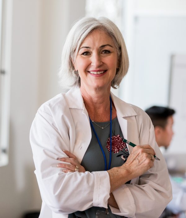 A smiling, mature woman in a lab coat stands in a bright office, holding a pen. Diverse colleagues are blurred in the background, suggesting a collaborative workspace.