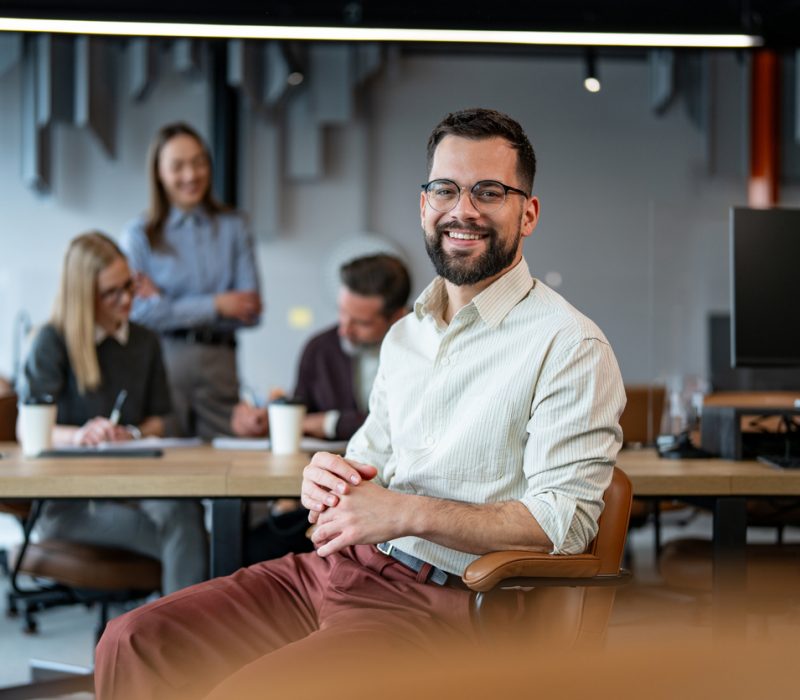 Smiling young adult businessman sitting in a modern corporate office, looking at the camera, with diverse colleagues collaborating at a meeting table in the blurred background